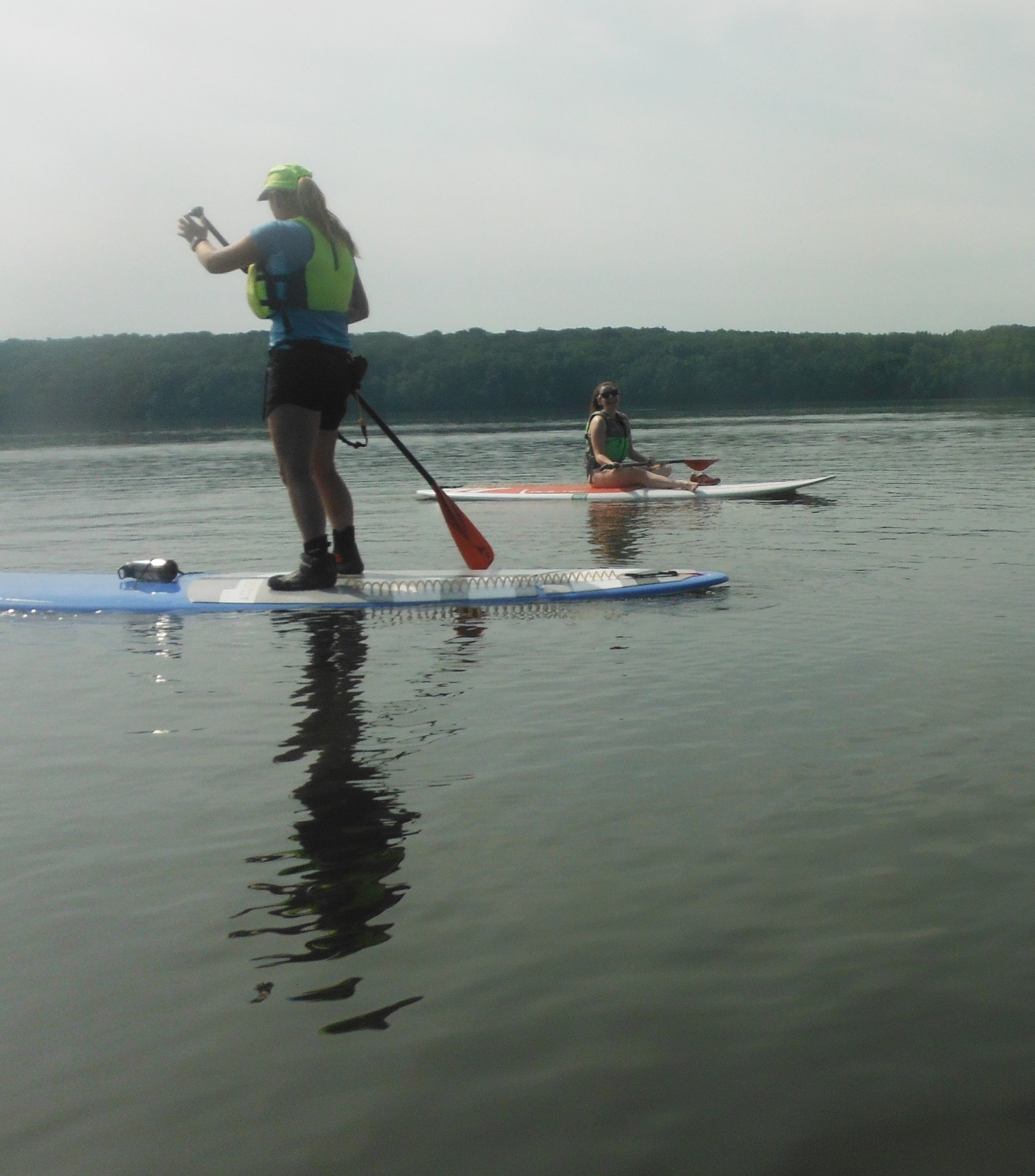 A woman on a stand up paddle board, another sits on one in the background