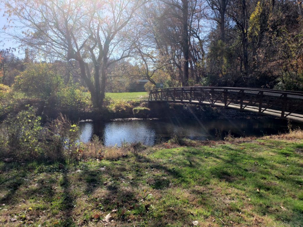 a creek with a walking bridge across it on the right side of the image. Both sides of hte creek have a grassy area