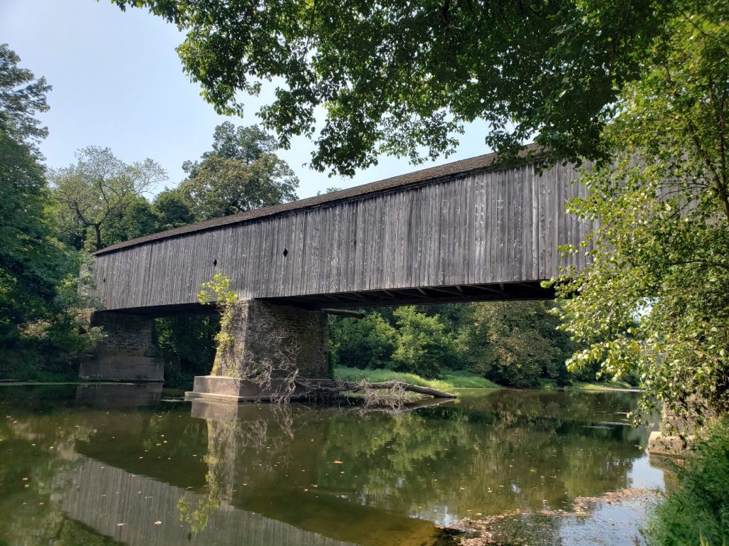 a weathered gray wooden covered bridge with stone supports over a creek; trees are on both banks