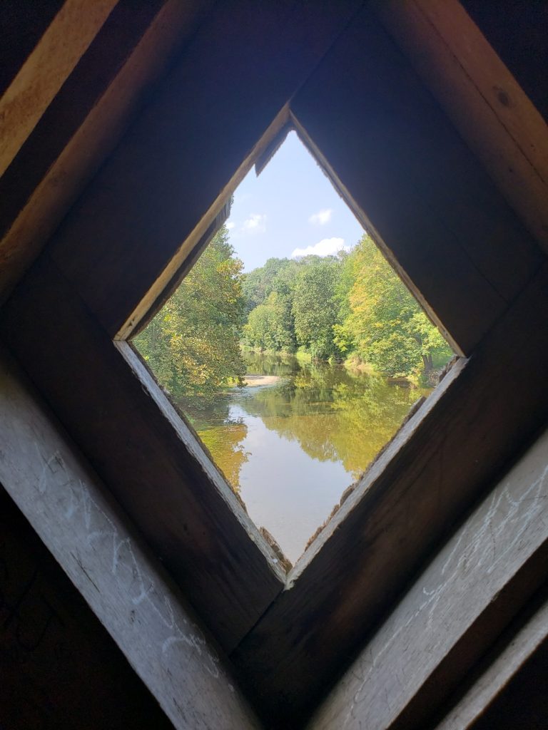 a diamond-shaped window in the bridge reveals a view of the creek which appears a greenish-brown; trees line the banks