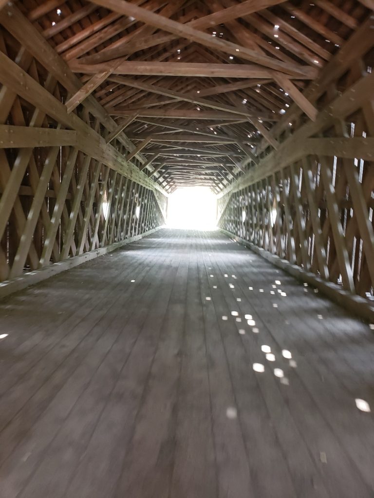 the interior of a covered bridge with exposed wood trusses; llight shines through small openings in the wood, creating dots of light on the wooden floor planks