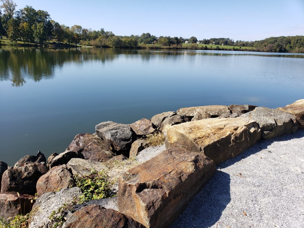 The view of Chambers Lake. A light-gray crushed gravel path leads to large rocks along the edge of the lake. Across the Lake, you can see deciduous trees reflecting in the blue water on the far side.